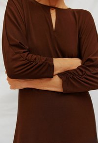 Braunes, langärmliges Kleid mit einem Schlüsselloch-Ausschnitt. Die Ärmel sind an den Manschetten gerafft und betonen einen glatten, strukturierten Stoff.