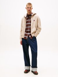 Felpa beige sovrapposta a una camicia a quadri rossa, jeans in denim blu scuro con risvolti e sneakers nere e rosse a fantasia. Design semplice e casual.