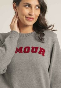 Pull en tricot gris avec le mot "AMOUR" en rouge en relief sur la poitrine. Encolure ronde et poignets côtelés ajoutent de la texture au design.