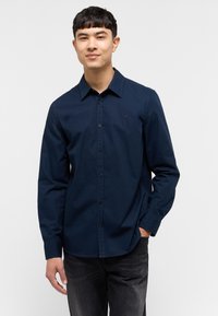 Camisa de botones azul marino de algodón con cuello apuntado, mangas largas y un pequeño logo en el pecho. Combinada con pantalones vaqueros oscuros.
