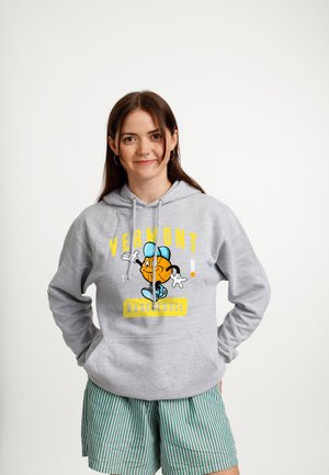 Jeune femme portant un sweat à capuche gris du basketball du Vermont avec un personnage de dessin animé en forme de ballon de basket et un short à rayures vertes, debout les mains dans les poches.