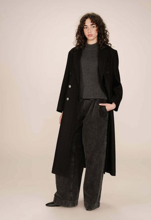 Femme aux cheveux bouclés portant un long manteau noir, un pull gris foncé, un jean noir à jambes larges et des chaussures noires à bouts pointus, se tenant avec une main dans la poche.