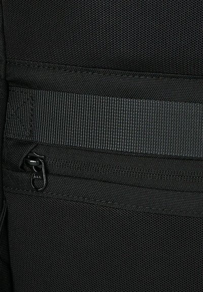 Tessuto nero testurizzato con una cintura orizzontale e una tasca con zip. Presenta un morbido tirante della zip con dettagli di cucitura.