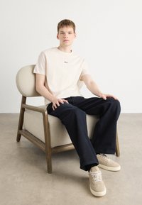 Lys beige bomull t-skjorte med liten svart logo, kombinert med mørkeblå vide bukser og beige sneakers, sittende på en trebjelle.