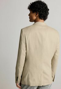 Blazer en lin beige avec une texture lisse, fente simple à l'arrière et accents de boutons noirs sur les poignets. Coupe ajustée, design structuré.