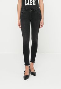 Μαύρα στενά τζιν με ξεθωριασμένη υφή, ξεφτισμένο τελείωμα και μπροστινές τσέπες. Συνδυασμένα με μαύρες μυτερές γόβες, φορεμένα με graphic τοπ χωρίς μανίκια.