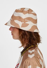 Brun och gräddfärgad bucket hat med vågig mönster, tillverkad av tyg. Har en logotyp på sidan och en bred brätte, bärs med en mönstrad skjorta.