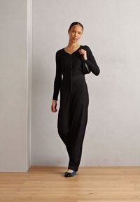 Maglietta nera a coste con maniche lunghe e zip, abbinata a pantaloni neri a vita alta. Semplici ballerine blu e una borsa nera completano l'outfit.
