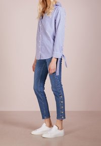 Camisa de botones a rayas azul y blanca con detalle de corbata en los puños, combinada con jeans ajustados azules que tienen acentos de botones en el dobladillo, y zapatillas blancas.