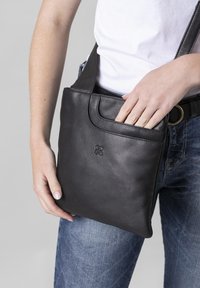 Black leather shoulder bag with a smooth texture, featuring a front pocket and an adjustable strap, held by a person wearing blue jeans.