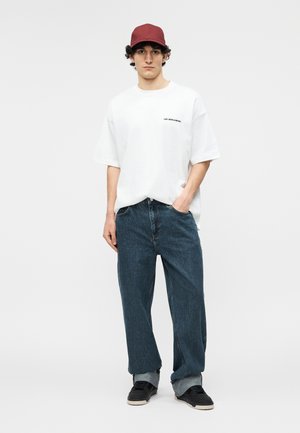 Fiatal férfi piros sapkával, fehér, túlméretezett "LES BENJAMINS" feliratú pólóban, bő kék farmernadrágban és fekete tornacipőben, egyik kezét a zsebében tartva áll.