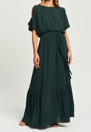 Frau trägt ein dunkelgrünes Maxikleid mit schulterfreien Ärmeln, tailliert mit einem Bindegürtel und gestuftem Saum, steht barfuß.