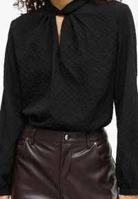 Svart teksturert bluse med nøkkelholldetalj ved halsen, kombinert med blanke, mørk burgunderrøde lærbukser med sidelommer.