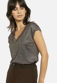 Top di bronzo lucido con scollo a V e maniche corte, tessuto leggermente strutturato, abbinato a pantaloni su misura marrone scuro. Effetto leggermente brillante.