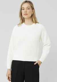 Pull en tricot côtelé blanc avec des manches longues et un col rond ; présente une coupe décontractée et un motif texturé, associé à un pantalon noir.