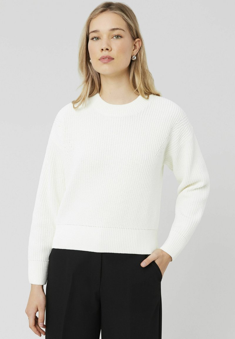 Pull en tricot côtelé blanc avec des manches longues et un col rond ; présente une coupe décontractée et un motif texturé, associé à un pantalon noir.