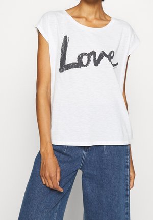 Vrouw draagt een wit mouwloos topje met dikke zwarte tekst "Love" en blauwe spijkerbroek, staand met één hand ontspannen langs haar zij.