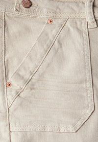 Bežová džínovina v detailu ukazuje přední kapsu se měděným nýtem a šitými detaily, zdůrazňující strukturovaný a odolný povrch.