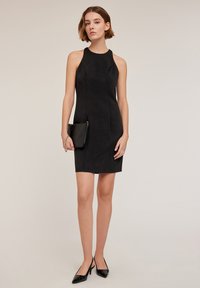 Svart ärmlös klänning med hög halsringning och figurnära silhuett, tillverkad av smidigt tyg, tillsammans med en liten svart handväska och spetsiga klackar.