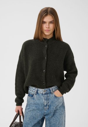 Giovane donna con lunghi capelli castani indossa un maglione verde scuro con bottoni e jeans chiari a vita alta, tenendo una borsa scura.