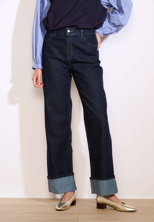 Jeans blu scuri a gamba larga con vita alta e orli arrotolati, abbinati a una blusa fluida a righe blu e bianche. Texture del denim liscia.
