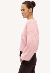 Pull rose duveteux avec un large décolleté, coupe ample et manches longues, associé à un pantalon marron foncé. Texture lisse avec une apparence douce.