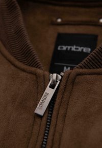 Brown suede zip-up jacket with ribbed collar, featuring a metal zipper pull with "Ombre" branding, and a label visible inside.