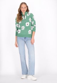 Grüner Strickpullover mit weißen Blumenmustern, mit einem Reißverschlusskragen, getragen mit hellblauen Jeans und weißen Sneakers. Das Model hat lange Haare, neutraler Hintergrund.