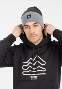 Grijze gebreide beanie met een donkergrijze bovenkant, voorzien van een zwart logo-label. De persoon draagt een zwarte hoodie met een witte bergafbeelding.