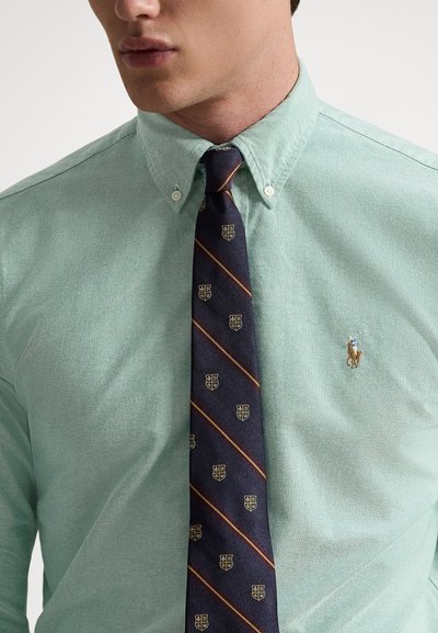 Šviesiai žalia marškiniai su tekstūruota medžiaga, derinami su tamsiai mėlynu kaklaraiščiu, puoštu įstrižomis juostelėmis ir skydo raštais. Ant marškinių mažas logotipas.