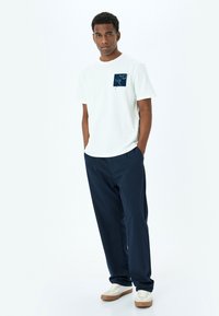Camiseta de algodón blanca con un bolsillo gráfico en azul marino, combinada con pantalones de pierna ancha en azul marino. El modelo lleva zapatillas blancas con suelas de goma color tawny.