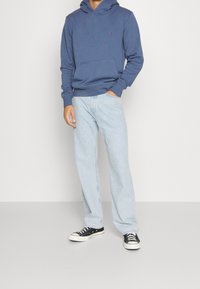 Hombre con sudadera azul, jeans de color azul claro y zapatillas negras, de pie con una mano en el bolsillo contra un fondo blanco.
