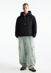 Giacca nera con cappuccio e tasca frontale, tessuto a trama; abbinata a pantaloni cargo oversize di un leggero blu e sneakers nere e gialle.