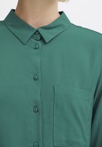 Groene button-up overhemd van gladde stof, met een puntige kraag, een borstzak en zes knopen in het midden met een eenvoudige uitstraling.