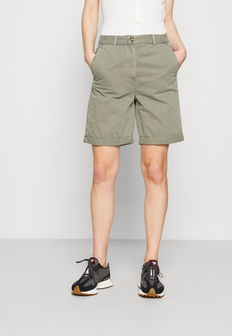 Personne portant un short vert olive jusqu'aux genoux, une chemise blanche et des chaussures de sport noires, debout les mains dans les poches sur un fond uni.