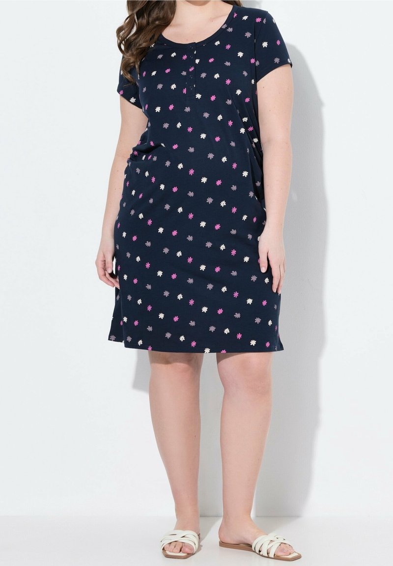 Femme portant une robe bleu marine mi-longue avec de petits motifs floraux roses, blancs et violets et des sandales blanches plates à enfiler.