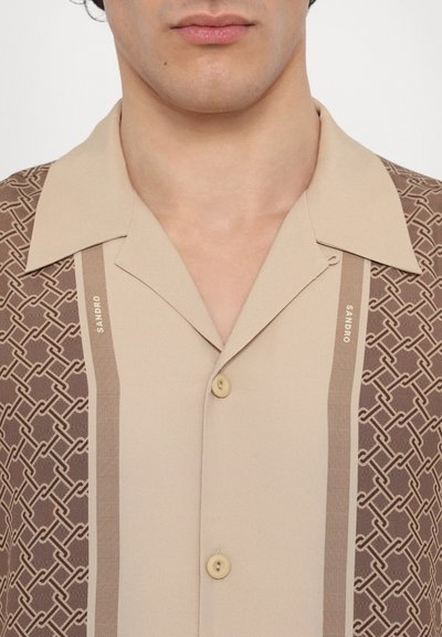 Chemise à manches courtes en tissu beige avec un motif géométrique marron. Elle présente un col et des boutons beiges, ainsi qu'un détail de logo.