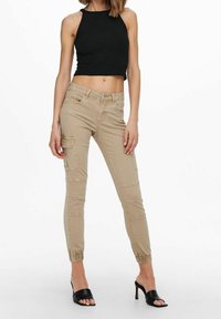 Pantalones cargo beige con puños elásticos en los tobillos, con bolsillos laterales y un diseño ajustado. Combinados con un top corto de canalé negro y tacones destalonados.