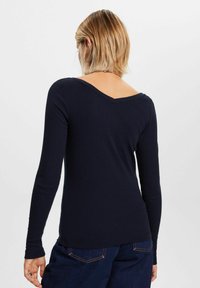 Laikkuva navy pitkähihainen paita V-muotoinen pääntie ja kapea muotoilu. Pehmeä rakenne ja viimeistelty helma. Malli kääntynyt pois päin, lyhyet hiukset.