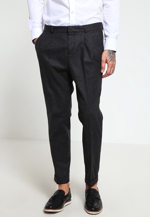 Man draagt donkergrijze taps toelopende broek met omslagen, een wit overhemd en zwarte leren loafers met kwastjes, op een lichte achtergrond.