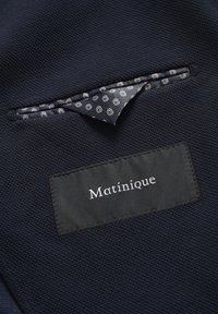 Sötét tengerészkék textúrájú anyag, kis zsebbel, mintás béléssel. Fekete címke, rajta a "Matinique" név fehér színnel hímezve.