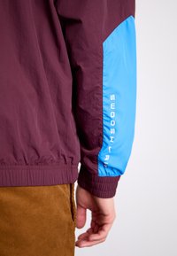 Bordeauxjacka med blå ärm som har texten "SWOOSH". Lättviktsmaterial, elastiska ärmslut och en rynkad fåll.