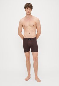 Καφέ αθλητικά σορτς με λεία υφή, ελαστική μέση και εφαρμοστό σχέδιο, εκτίθενται σε άνδρα μοντέλο που στέκεται μπροστά από λευκό φόντο.