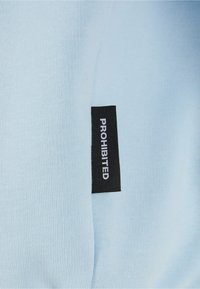 Helles blaues Tuch mit glatter Textur, versehen mit einem schwarzen Etikett, auf dem "VERBOTEN" steht. Der Stoff hängt, was seine weiche Qualität zeigt.