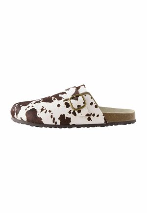 Braun-weiß gefleckte Clogs mit flacher Sohle, verstellbarem Riemen und Metall-Schnalle. Weiche Textur, geschwungene Form und minimales Profil.