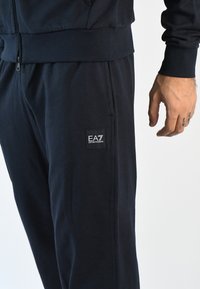 Conjunto de chándal azul marino hecho de tejido suave, que incluye una chaqueta con cremallera, puños acanalados y un pequeño parche cuadrado con logo en la pierna del pantalón.