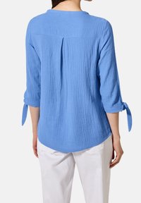 Femme portant un chemisier bleu clair texturé à manches 3/4 nouées aux poignets, associé à un pantalon blanc, vue de dos.