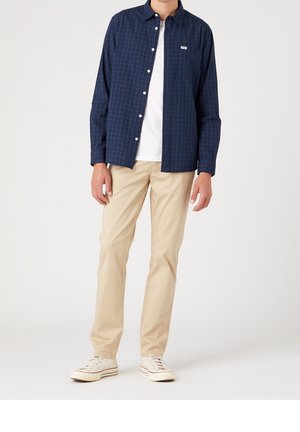 Uomo che indossa pantaloni beige, sneaker bianche, t-shirt bianca e camicia a quadri blu navy con logo Wrangler sulla tasca del petto.