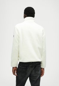 Pull en polaire blanche avec un col haut, présentant une surface texturée. Associé à un jean en denim foncé, le design est simple et fonctionnel.