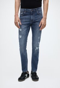 Modré džínové kalhoty s úzkým střihem, zdobené ošoupanými záplatami na přední části a klasickým pětiprázdným designem. Nošeno s černými teniskami.